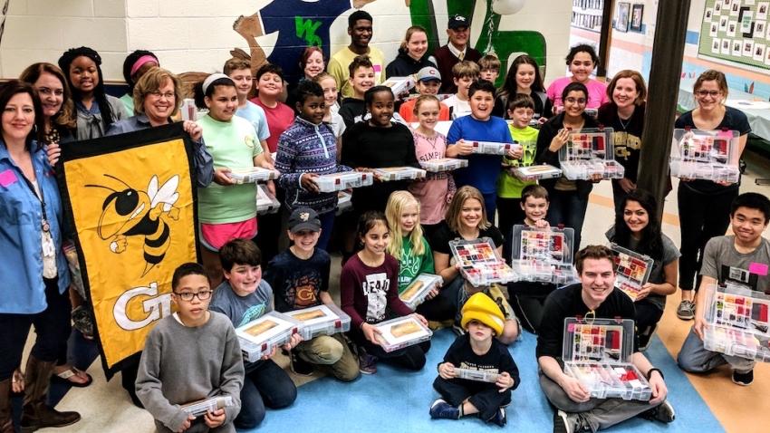 Group photo of Georgia Tech Computing students and faculty and elementary students in school hallway with Georgia Tech Buzz poster at left 