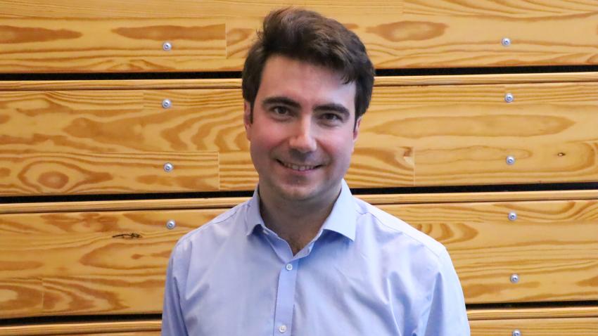 A man standing in front of a wooden wall. He is smiling and tilting his head. 