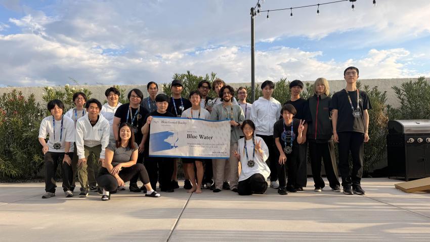 A group of people standing outside holding a banner. The banner says "Blue Water" which is the team's name.