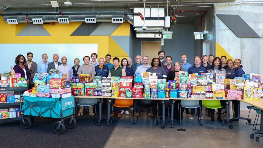 1.A photo of a group of people standing behind a table full of packaged food. The group is smiling and represents a diverse crowd of faculty and staff.