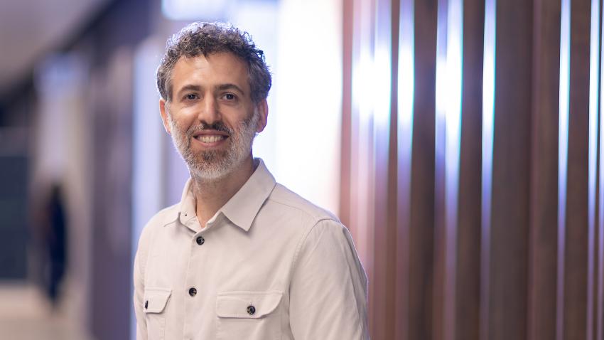 A man with salt and pepper hair and beard stands in a hallway wearing a white buton up shirt. There is a modern wooden panel behind him which reflects light and the purple color from the other walls.