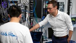 A man points to a rack of computer monitors. Another man sits in front of a laptop with his back to the camera. 
