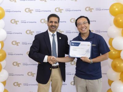 Two men shaking hands. One is holding a certificate. They are standing in front of a step and repest banner.