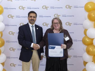 A man and a woman shake hands in front of a step and repeat banner. The woman is holding a certificate.