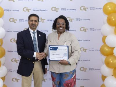 A man and a woman shake hands in front of a step and repeat banner. The woman is holding a certificate.