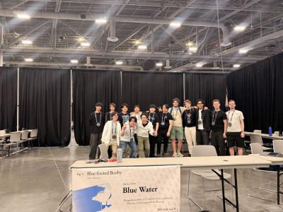 A group of people standing inside of a convention hall. Their banner has their team name, Blue Water