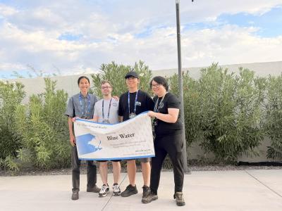 A group of four people stand outside holding up a banner. The banner has their team name, Blue Water