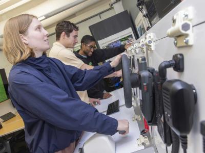 A group of students working on lab equipment. The equipment replicates a ship's helm. 